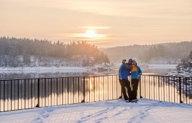 Die sanften H&uuml;gel des Waldviertels sind in eine glitzernde Schneedecke geh&uuml;llt, w&auml;hrend die Sonne langsam am Horizont aufgeht. Zwei Wanderer genie&szlig;en den atemberaubenden Ausblick auf den Stausee Ottenstein und die umliegende Winterlandschaft, die in warmen Gold- und Oranget&ouml;nen erstrahlt. Ein perfekter Moment, um die Ruhe und Sch&ouml;nheit der Natur zu erleben.