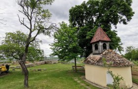 Kleine Kapelle mit rotem Ziegeldach in einer gr&uuml;nen Landschaft, umgeben von B&auml;umen und B&auml;nken.