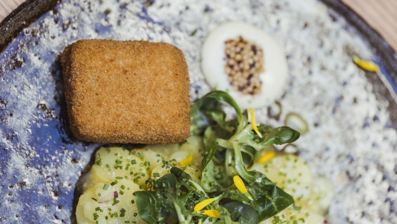 Ein Teller mit paniertem Kalbskopf, Kartoffelsalat und Feldsalat, garniert mit gelben Bl&uuml;ten.