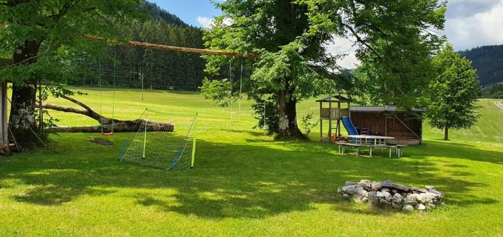 Ein Spielplatz auf einer grünen Wiese mit Schaukeln, einem Klettergerüst, einer Rutsche und einer Feuerstelle. Im Hintergrund sind Bäume und Hügel zu sehen.