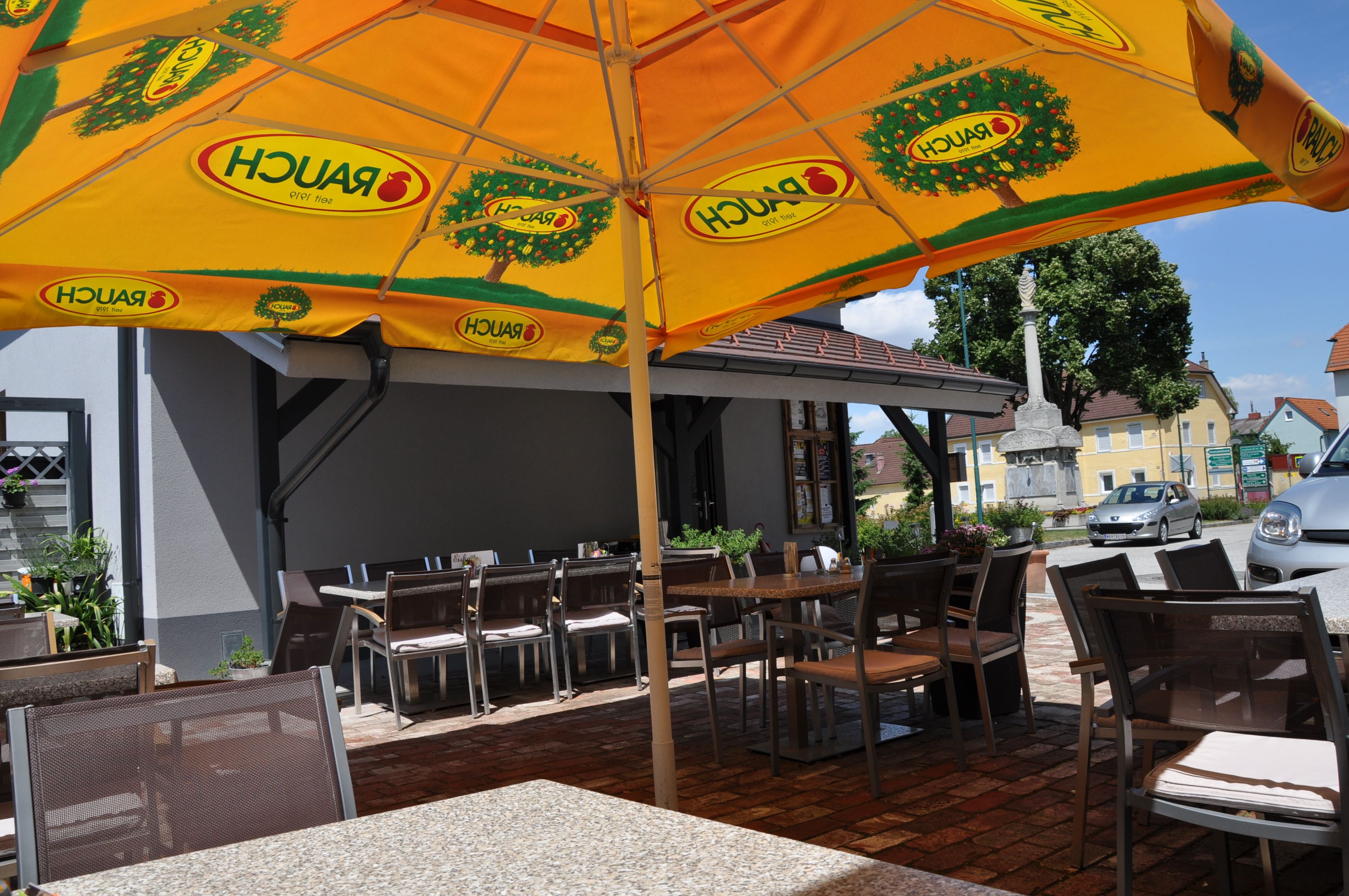 Außenbereich eines Cafés mit gelbem Sonnenschirm und leeren Tischen und Stühlen.