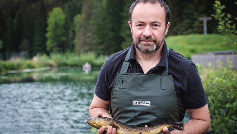 Ein Mann in gr&uuml;ner Sch&uuml;rze h&auml;lt einen Fisch vor einem Teich.