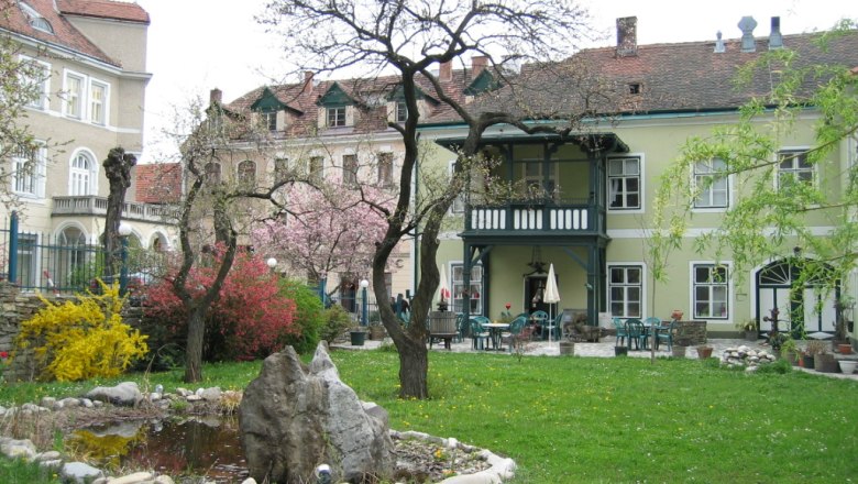 Ein Garten mit Teich und Bäumen vor einem historischen Gebäude mit Balkon.