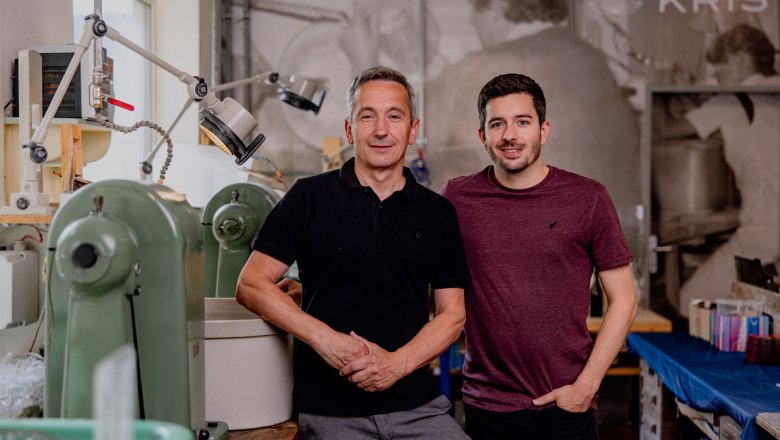 Zwei Männer stehen in einer Werkstatt mit Maschinen im Hintergrund.