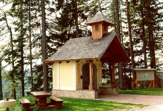 Kleine Kapelle im Wald mit Holztisch und Bank daneben.