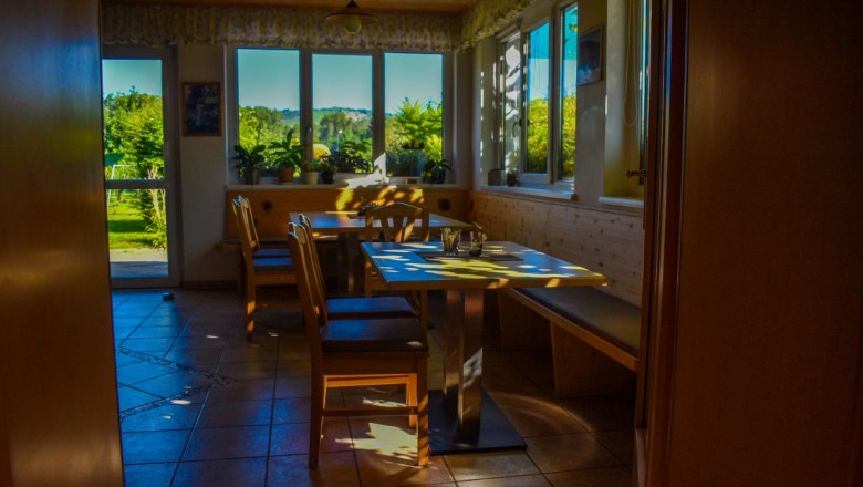 Ein gemütlicher Frühstücksraum mit Holzmöbeln und großen Fenstern, durch die Sonnenlicht fällt.