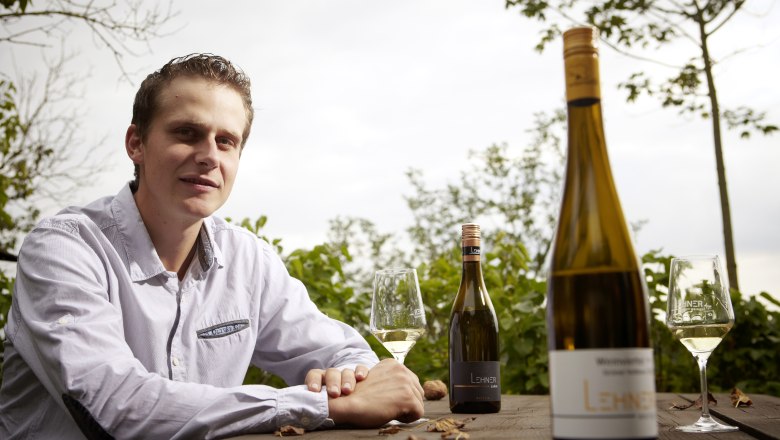 Ein Mann sitzt an einem Holztisch im Freien mit Weinflaschen und Gl&auml;sern.