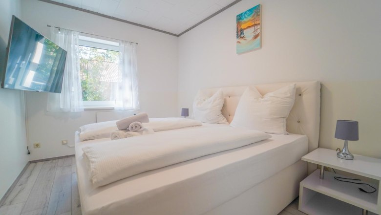 Modernes Schlafzimmer mit Doppelbett, weißer Bettwäsche, Wandbild, Fenster mit Vorhängen und Flachbildfernseher.