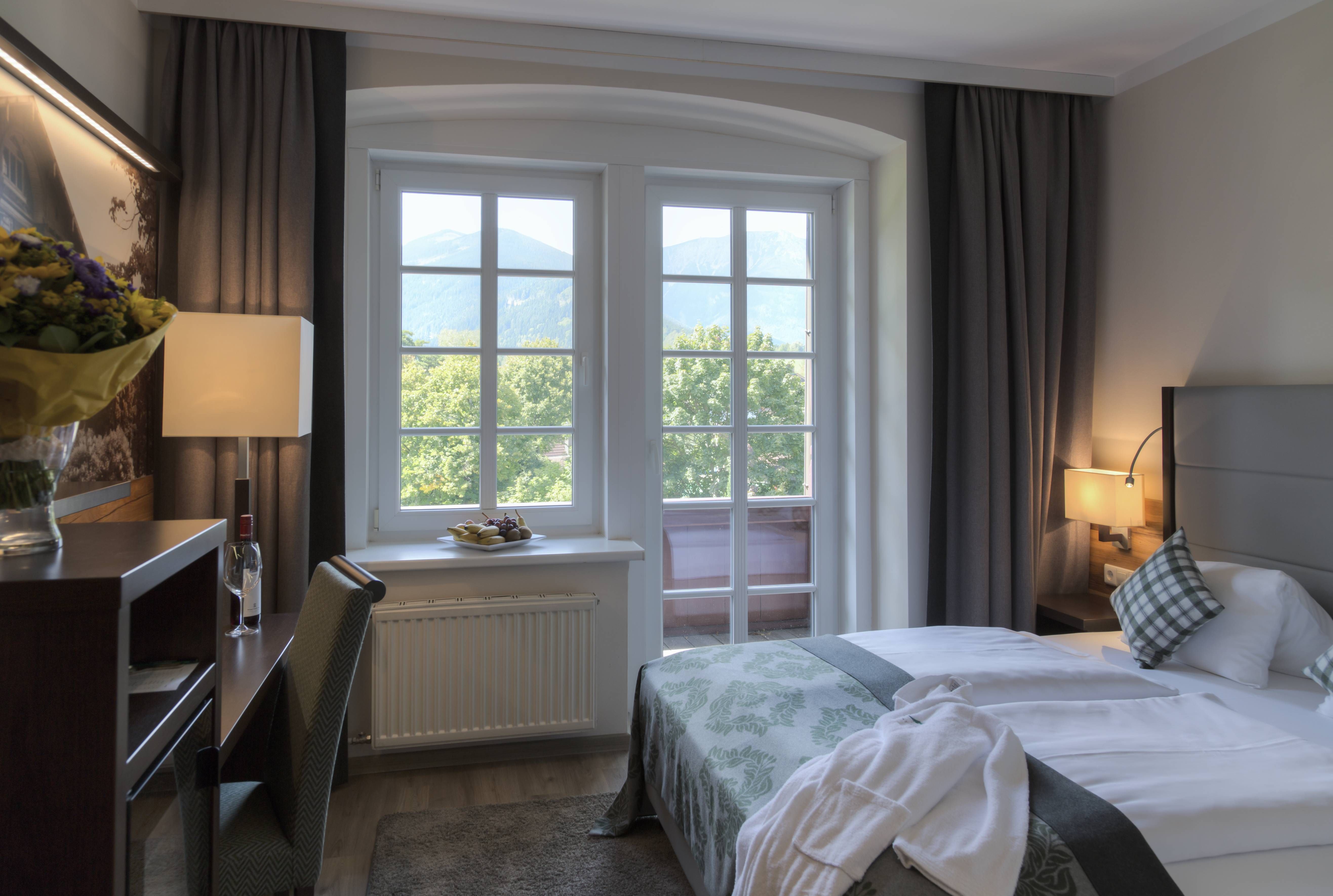 Ein gemütliches Hotelzimmer mit Doppelbett, Fenster mit Bergblick, Schreibtisch und Blumen.