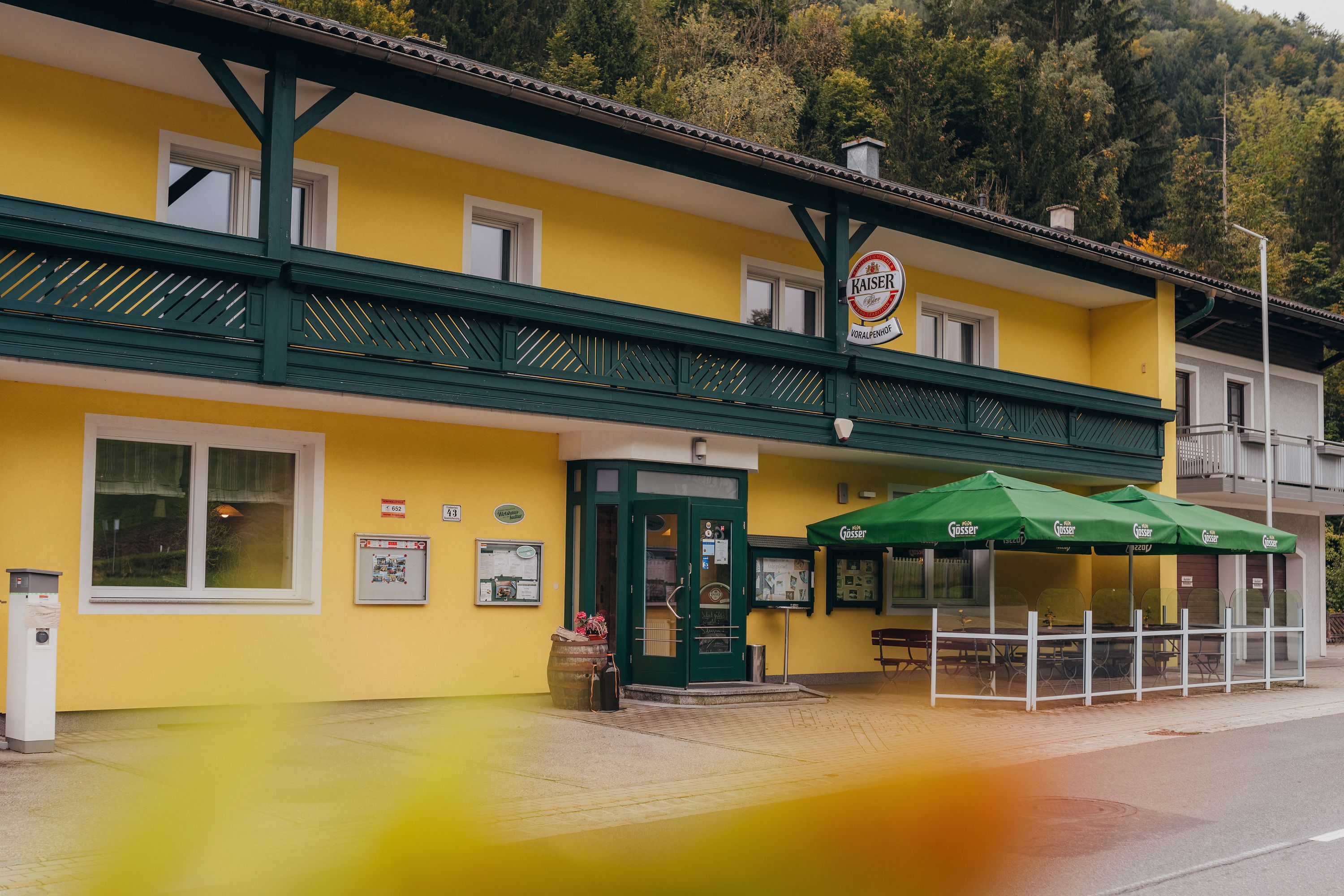 Gelbes Wirtshaus mit grünen Markisen und Schildern, umgeben von Bäumen.