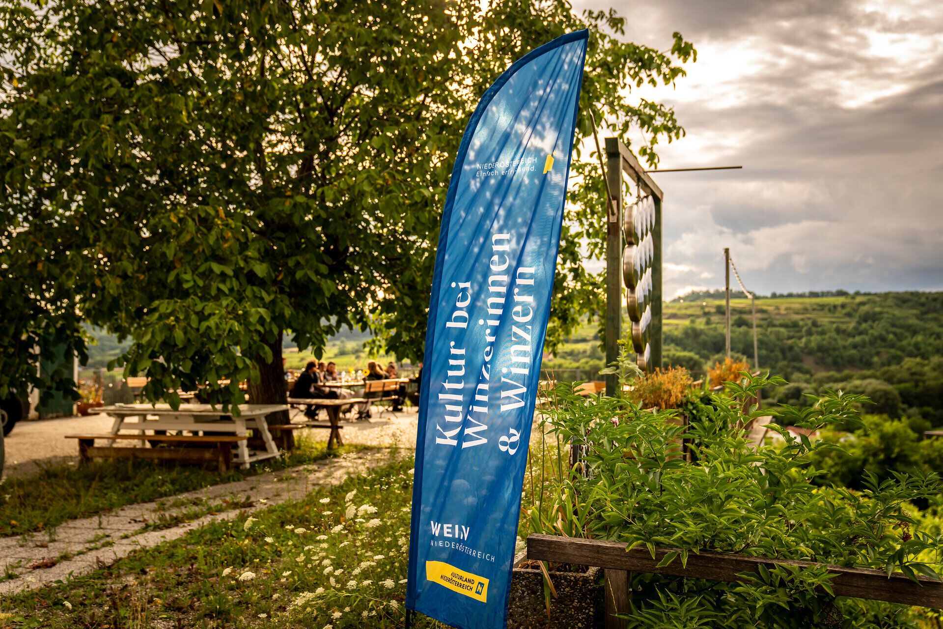 Blauer Banner „Kultur bei Winzerinnen & Winzern“ im Garten eines Weinguts. Im Hintergrund sitzen Gäste an Holztischen unter Bäumen, mit Blick auf die Weinlandschaft.