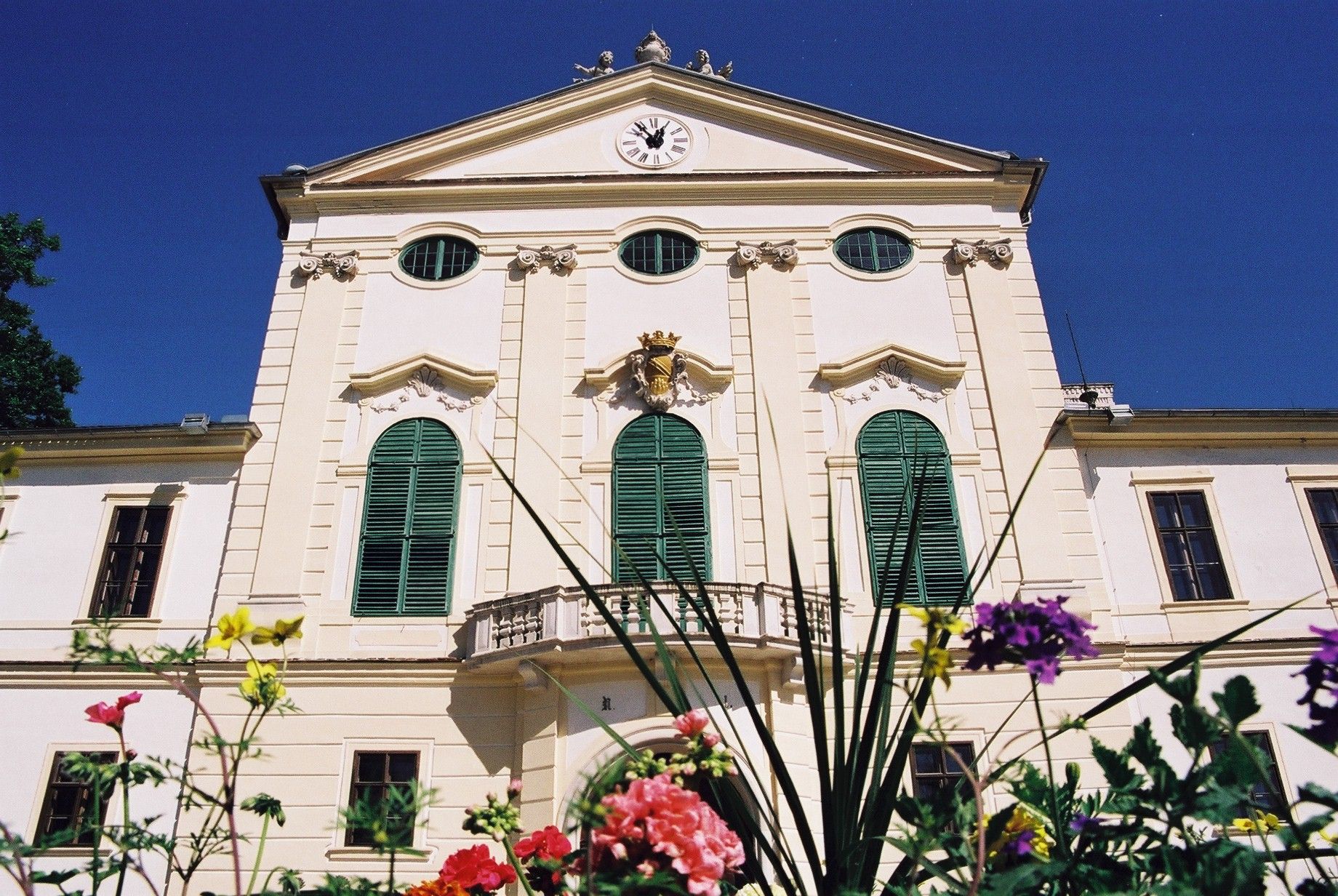 Fassade von Schloss Kirchstetten mit grünen Fensterläden und Uhr, umgeben von bunten Blumen.