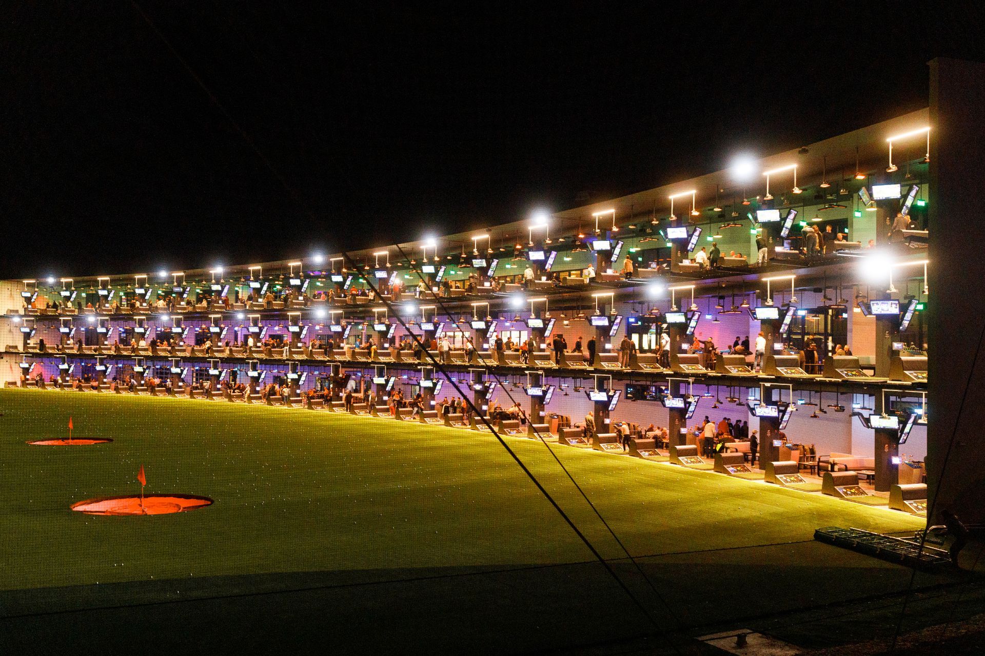 Beeindruckende Abendstimmung bei Topgolf mit beleuchteten Abschlagplätzen auf mehreren Ebenen und zahlreichen Gästen beim gemeinsamen Spielen und Genießen.