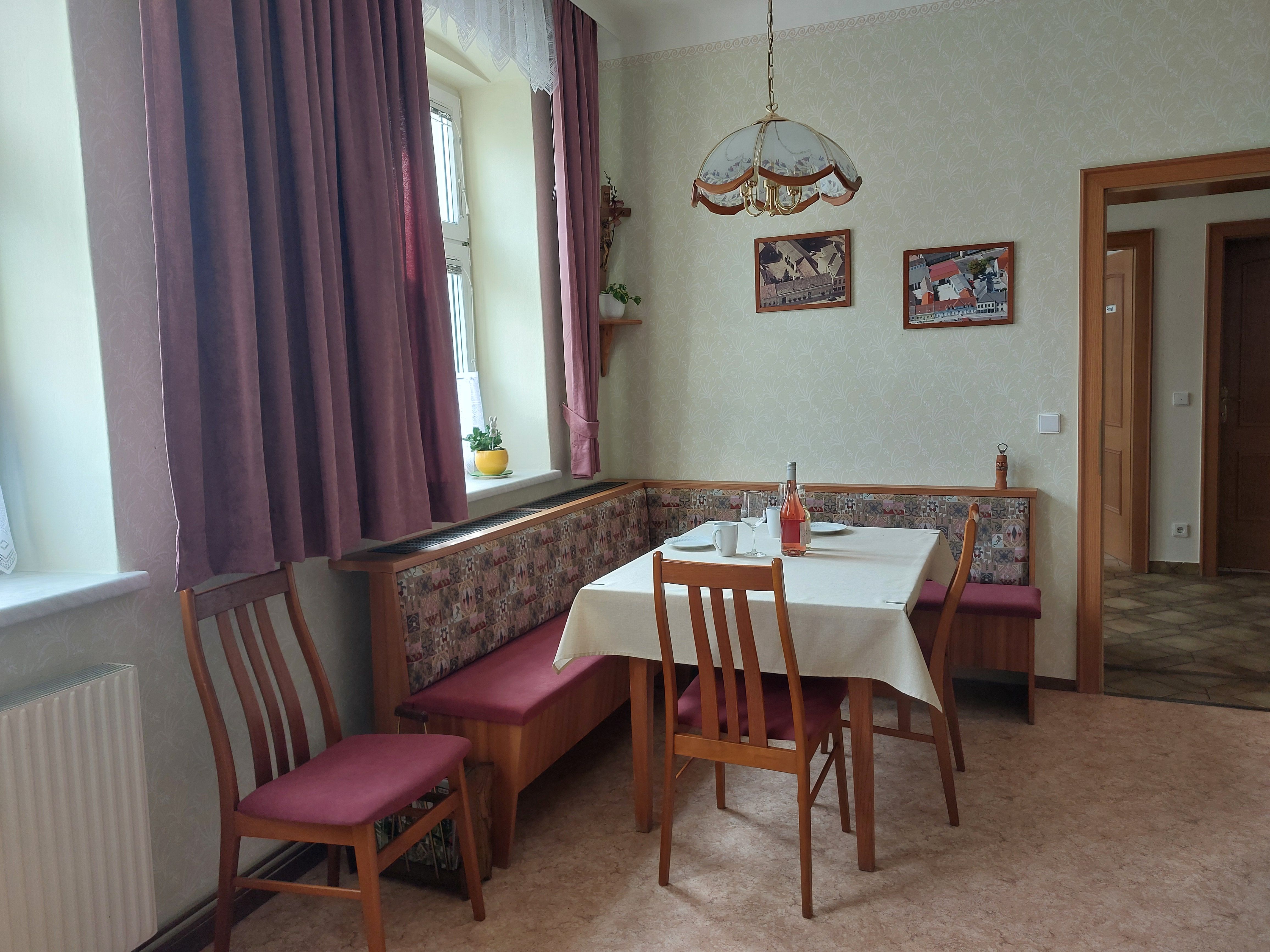 Gemütliche Wohnküche mit Esstisch, Stühlen und Eckbank. Fenster mit Vorhängen, Bilder an der Wand.