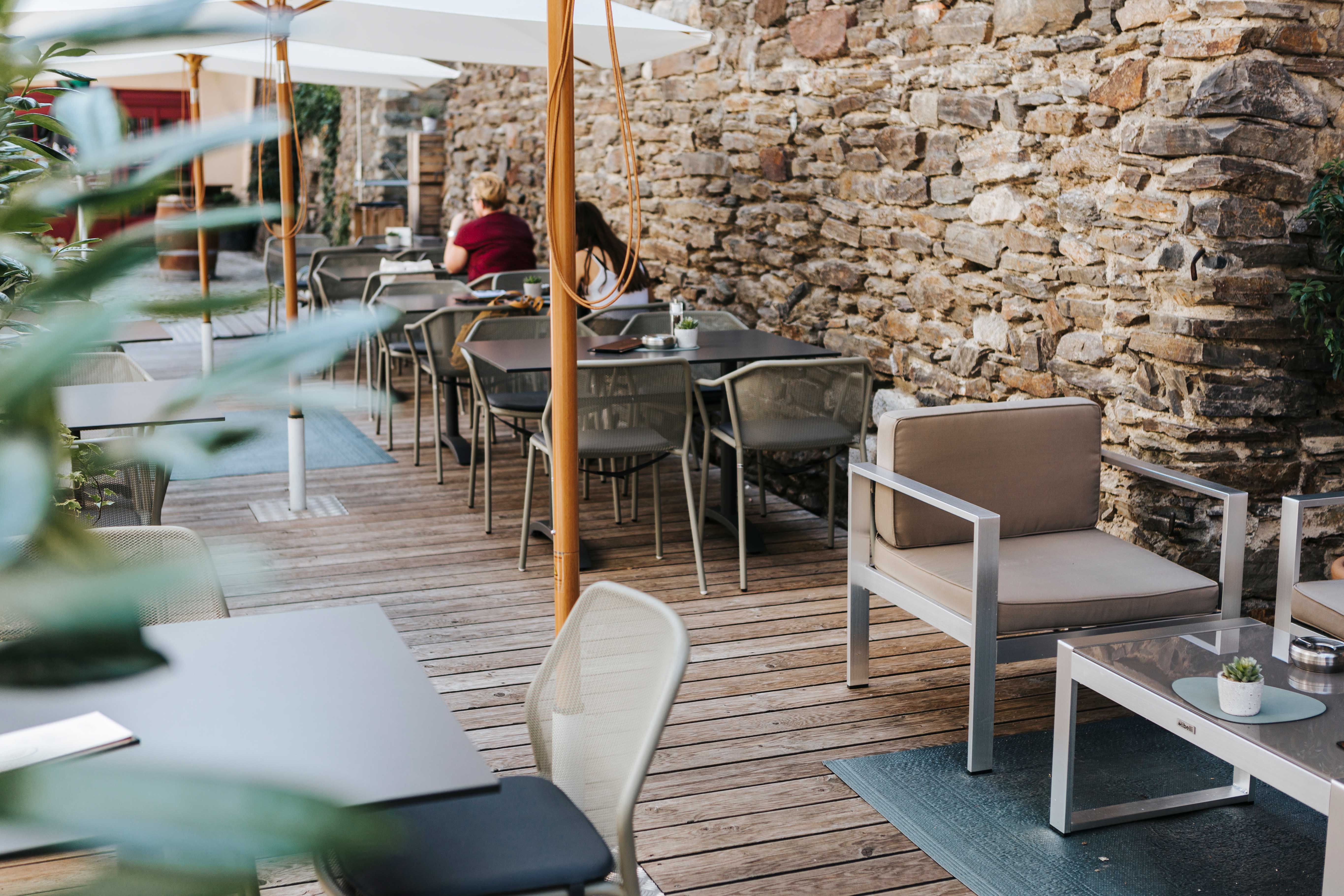 Gemütlicher Außenbereich eines Cafés mit Holzboden, modernen Möbeln und Steinmauer.