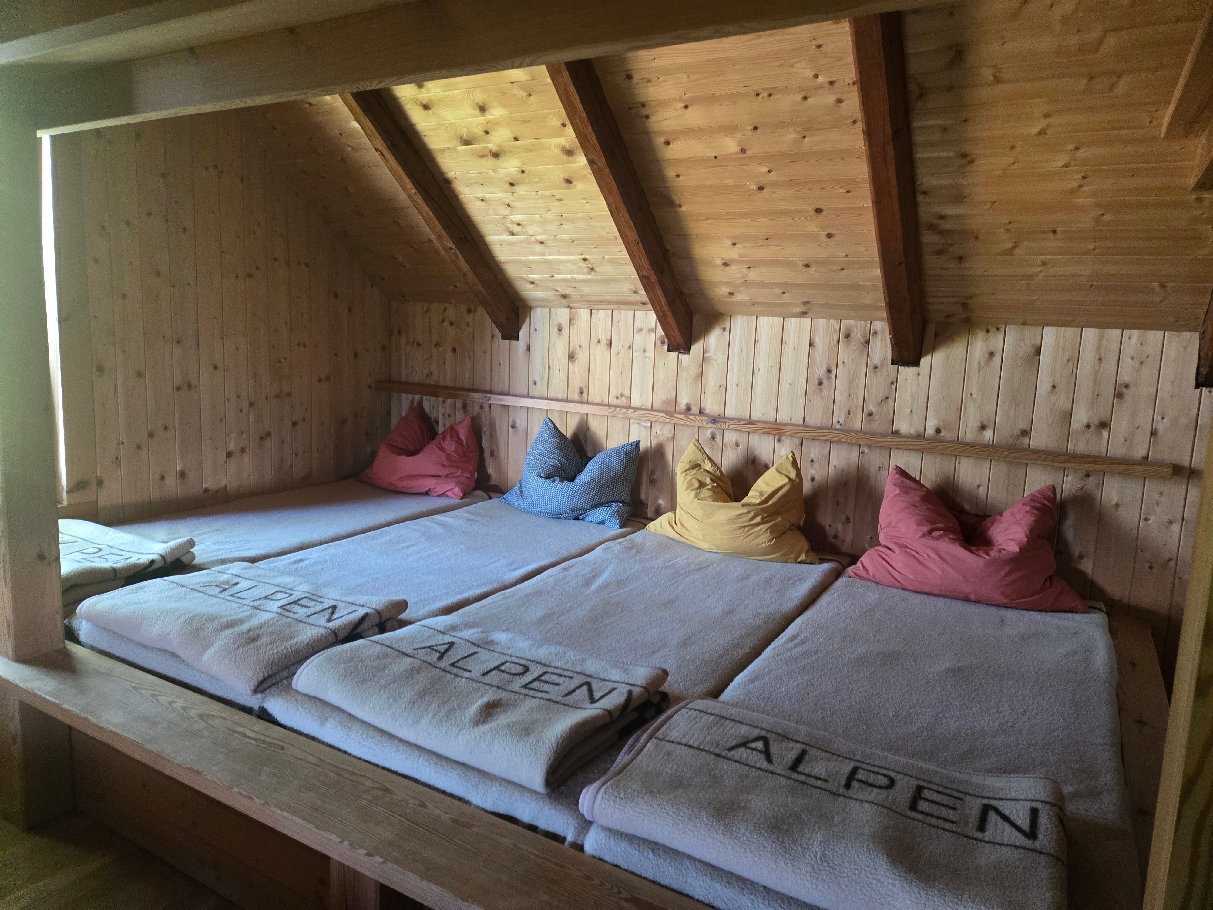 Innenraum der Enzianhütte mit vier Lagerplätzen unter einer Holzschräge, Decken und farbigen Kissen auf den Betten.
