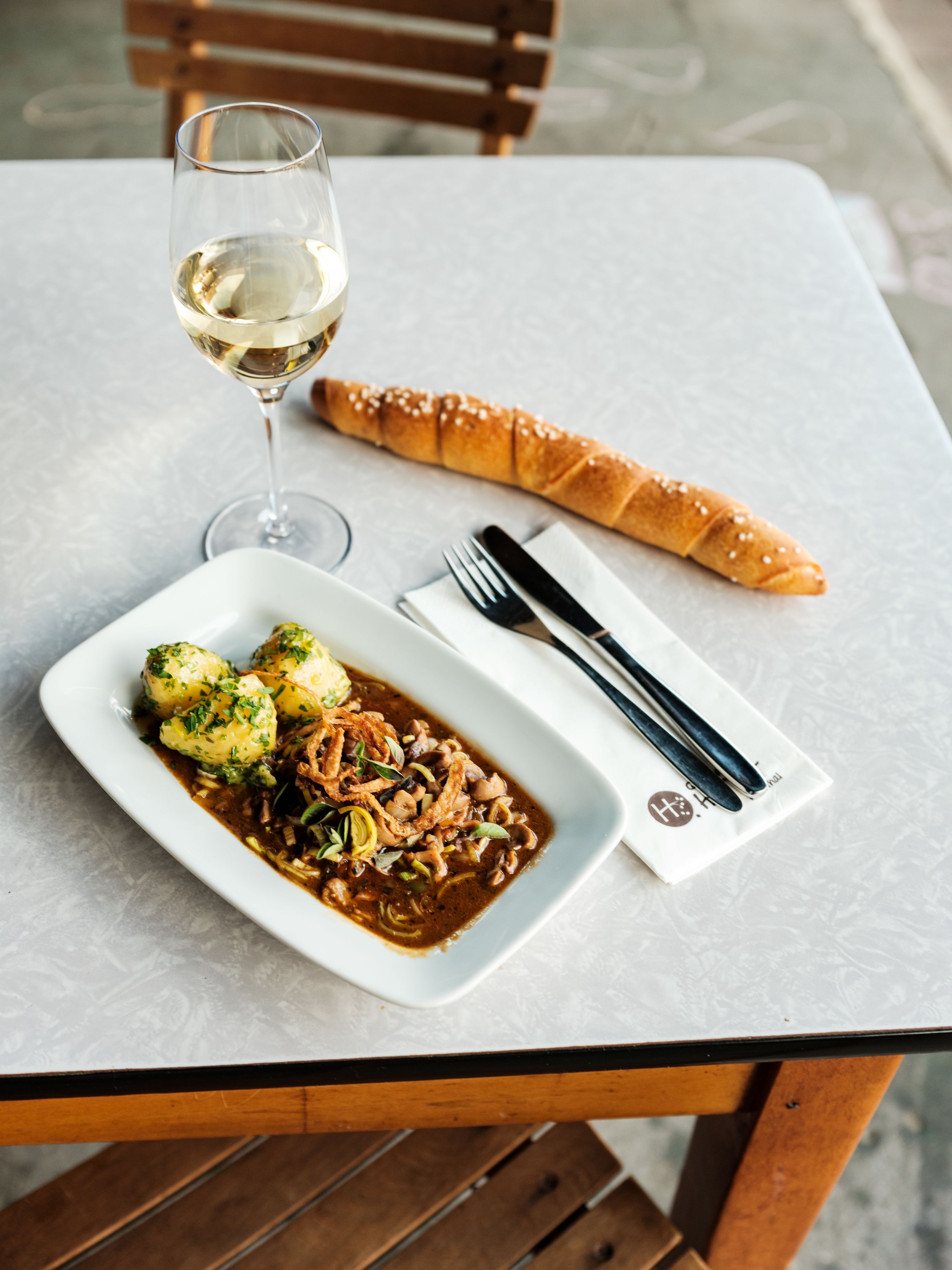 Ein Tisch mit einem Glas Weißwein, einem Teller mit Fleischgericht und Knödeln, einem langen Brötchen und Besteck auf einer Serviette.