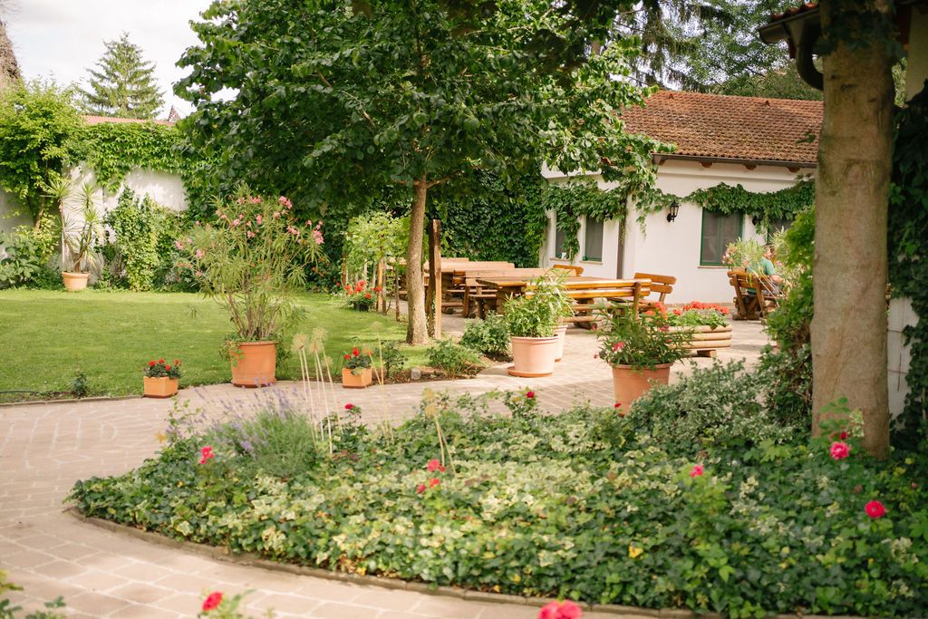 Ein gepflegter Garten mit Rasen, Blumenbeeten und Terrakotta-Töpfen. Im Hintergrund ein Haus mit Terrasse und Holztischen.