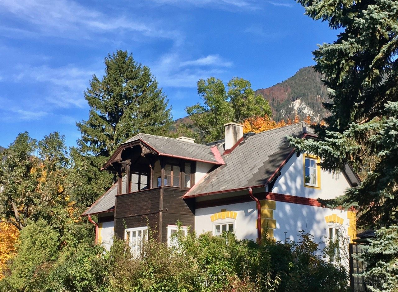 Ein malerisches Landhaus mit grauem Dach und gelben Fensterrahmen, umgeben von Bäumen und Bergen im Hintergrund.