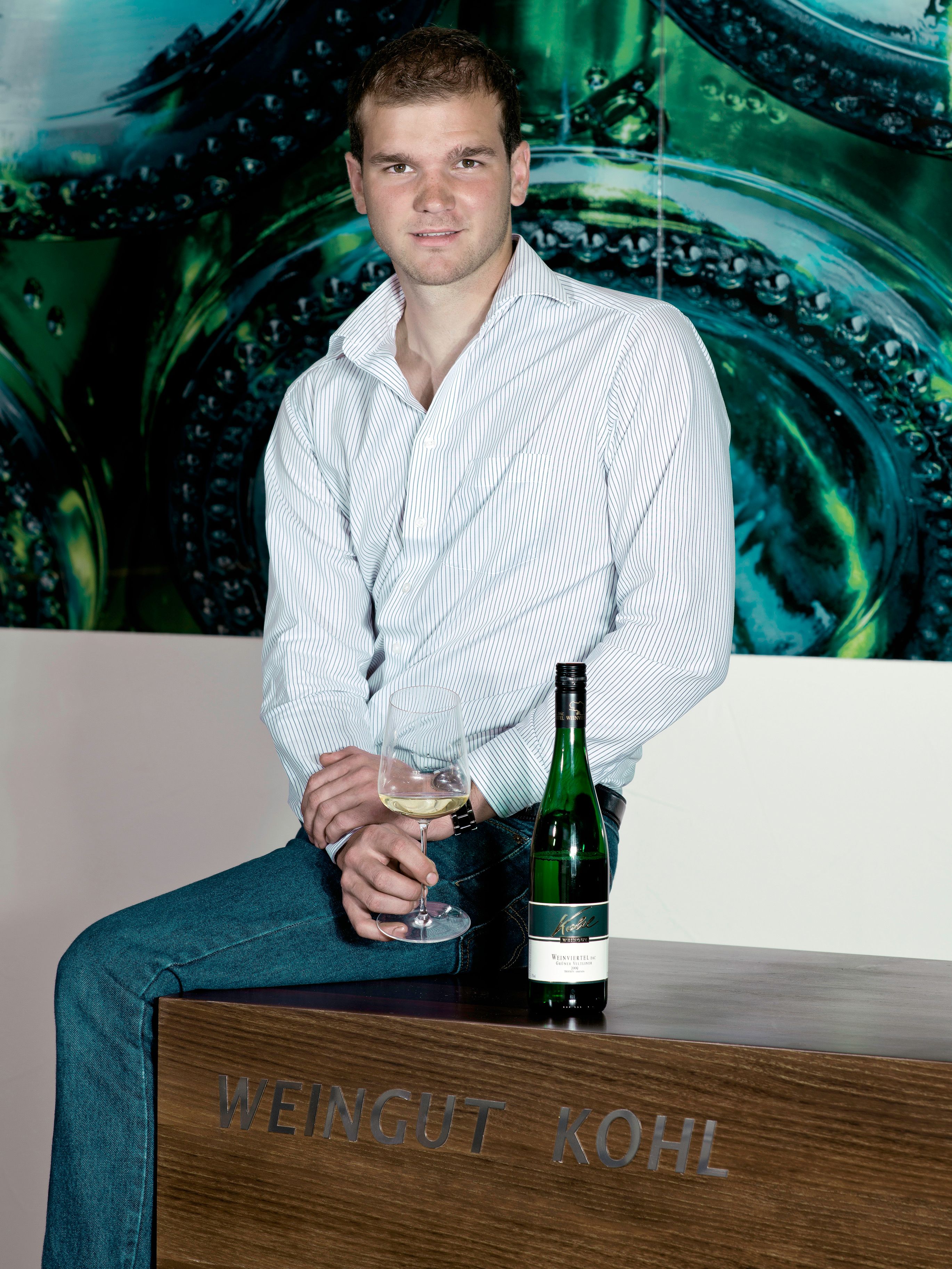 Ein Mann sitzt auf einem Tisch mit der Aufschrift 'Weingut Kohl', hält ein Weinglas und eine Weinflasche steht neben ihm.