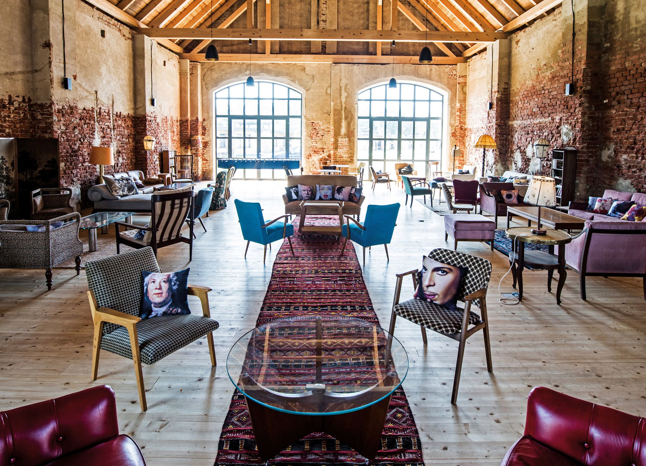 Innenansicht einer stilvoll eingerichteten Scheune mit Holzboden, gemauerten Wänden und großen Fenstern. Verschiedene Sitzmöbel und bunte Kissen sind zu sehen.