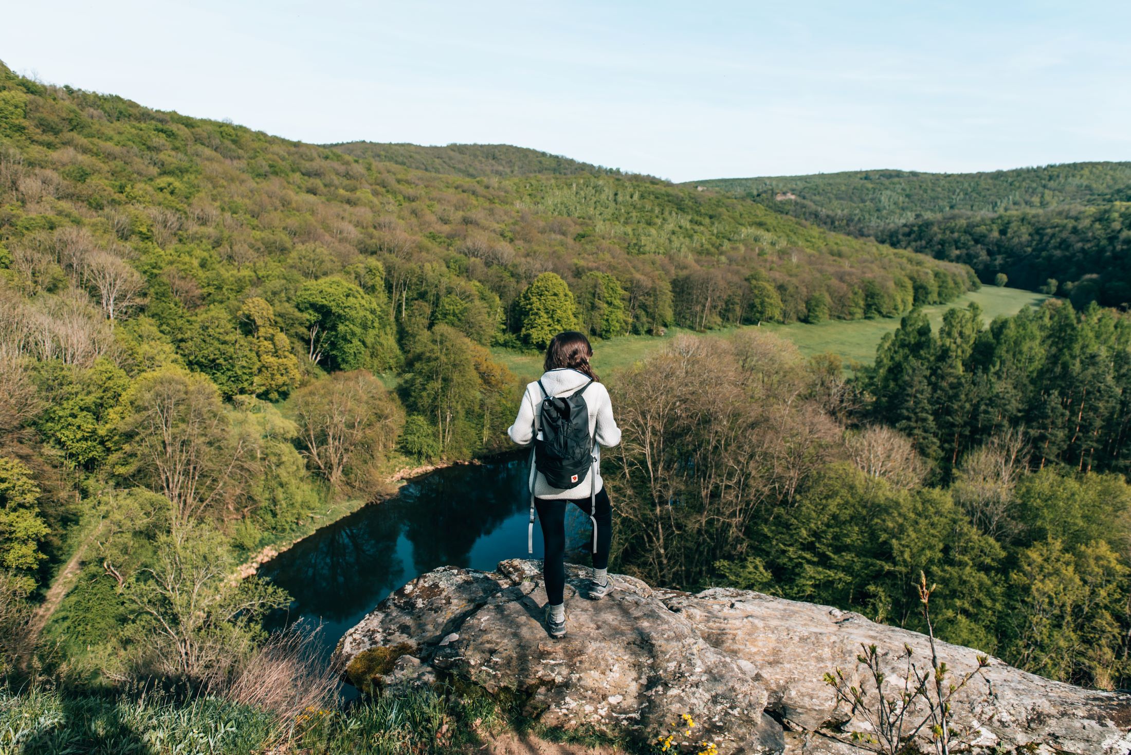 Person mit Rucksack steht auf einem Felsen und blickt auf bewaldete Landschaft und Fluss.