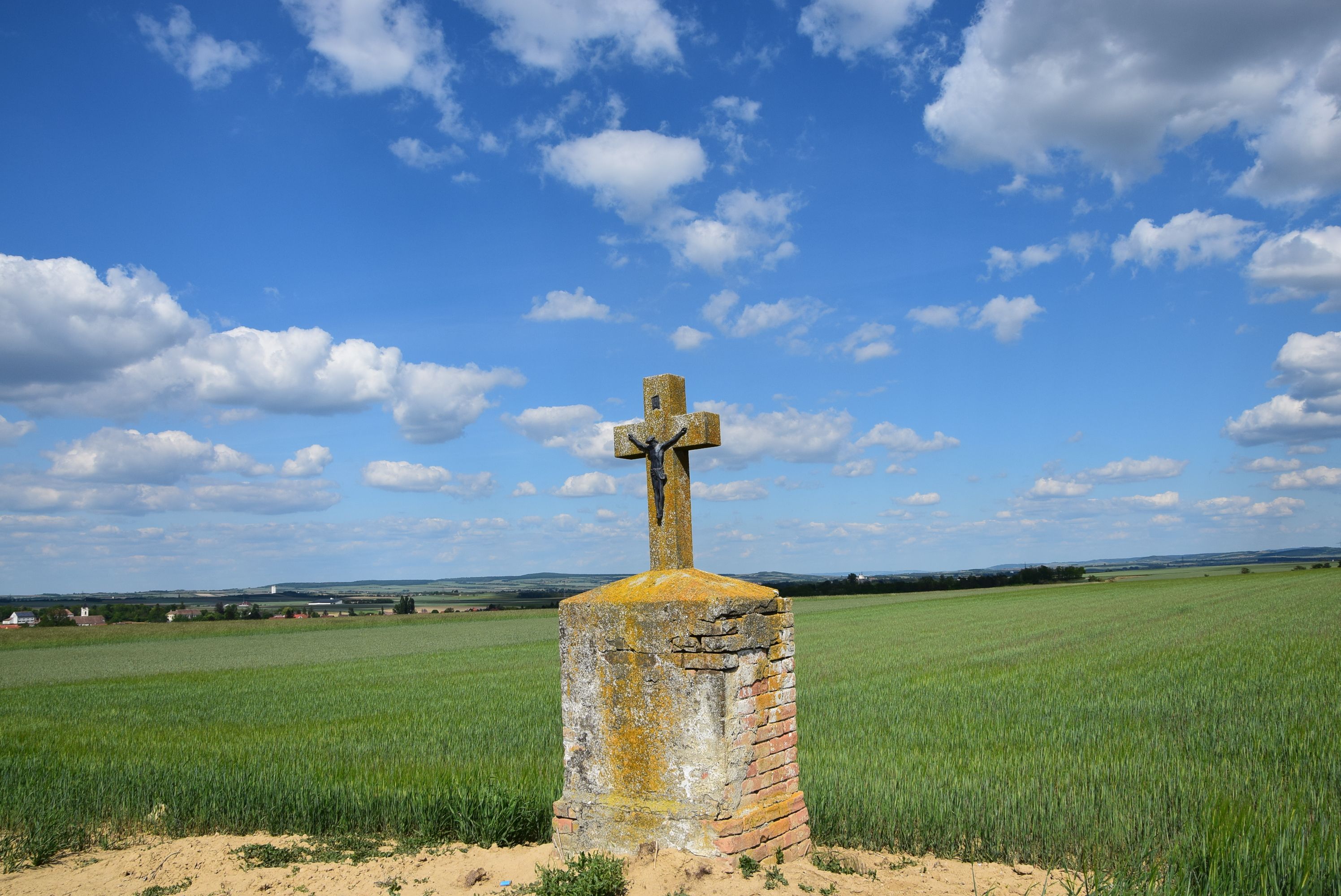 Ein steinernes Kreuz mit Christusfigur steht auf einem Feld unter blauem Himmel mit Wolken.