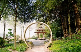 Ein orientalischer Garten mit einem runden Torbogen und einer Pagode im Hintergrund, umgeben von Bäumen und Pflanzen.