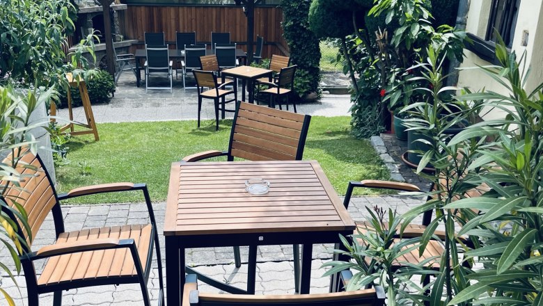 Ein gemütlicher Gastgarten mit Holztischen und -stühlen, umgeben von üppigem Grün und Pflanzen.