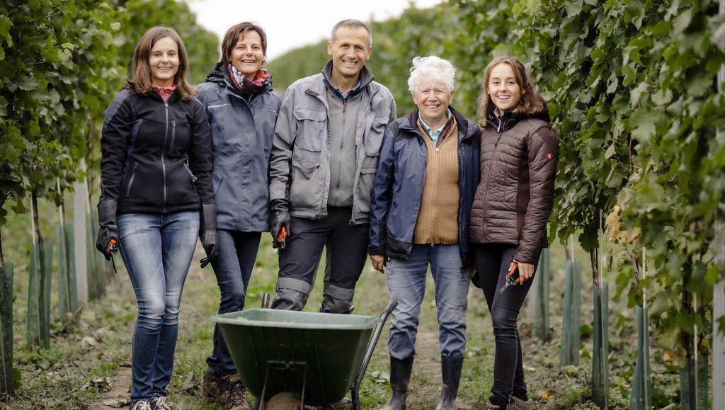 Fünf Personen stehen lächelnd in einem Weinberg mit einer Schubkarre.