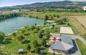 Luftaufnahme eines Badeteichs mit Liegewiese, B&auml;umen und einem Geb&auml;ude mit Sonnenschirmen.