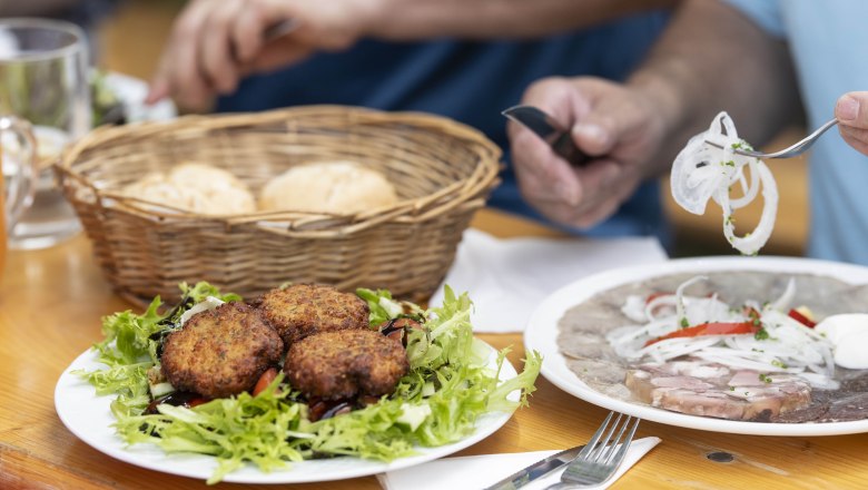Ein Tisch mit einem Teller voller Fleischlaberl auf Salat, einem Korb mit Geb&auml;ck und einem Teller mit Presswurst und Zwiebeln.