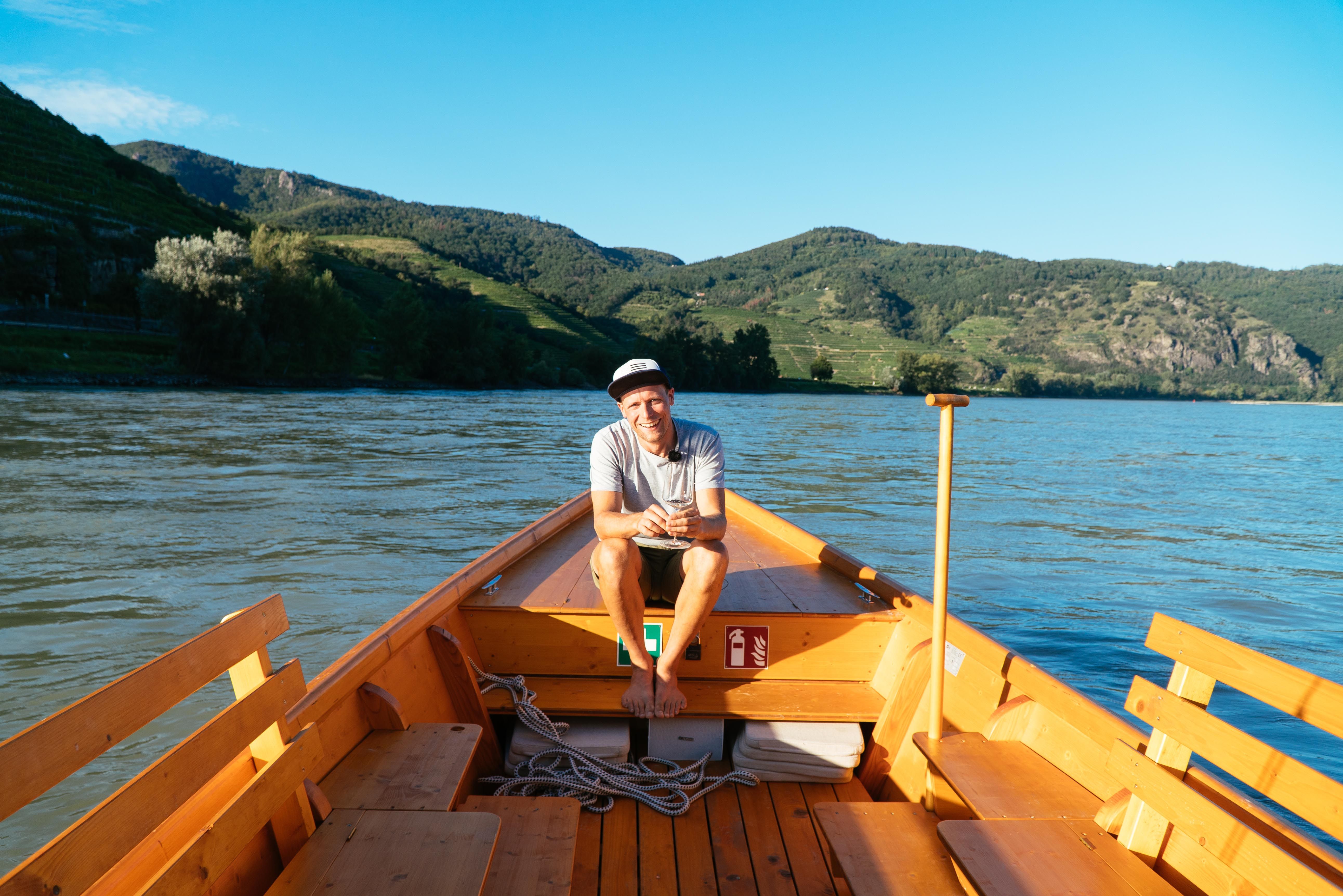 Person sitzt auf einem Holzboot auf einem Fluss, umgeben von grünen Hügeln.