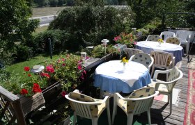 Terrasse mit Tischen und St&uuml;hlen, umgeben von Pflanzen und Blumen, mit Blick auf einen Fluss.