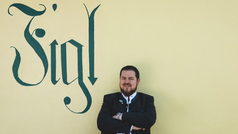 Ein Mann in traditioneller Kleidung steht vor einer gelben Wand mit dem Schriftzug 'Figl'.