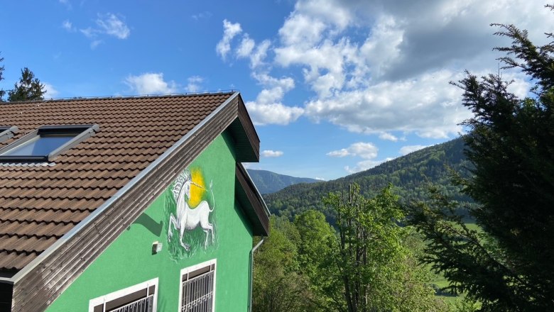 Gr&uuml;nes Haus mit Einhornbild an der Wand, umgeben von B&auml;umen und Bergen unter blauem Himmel.
