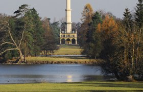 Ein Minarett im Schlosspark von Lednice, umgeben von Bäumen und einem See im Vordergrund.