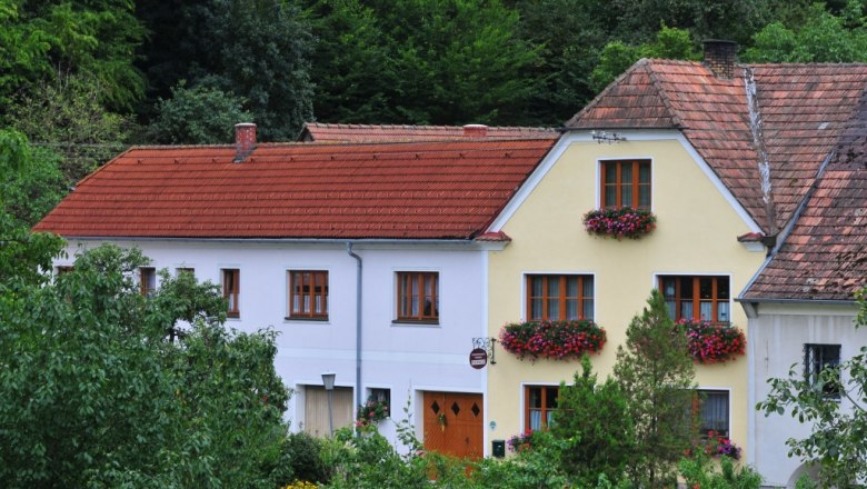 Ein traditionelles Haus mit roten Dachziegeln und bl&uuml;henden Blumenk&auml;sten vor den Fenstern, umgeben von gr&uuml;ner Vegetation.