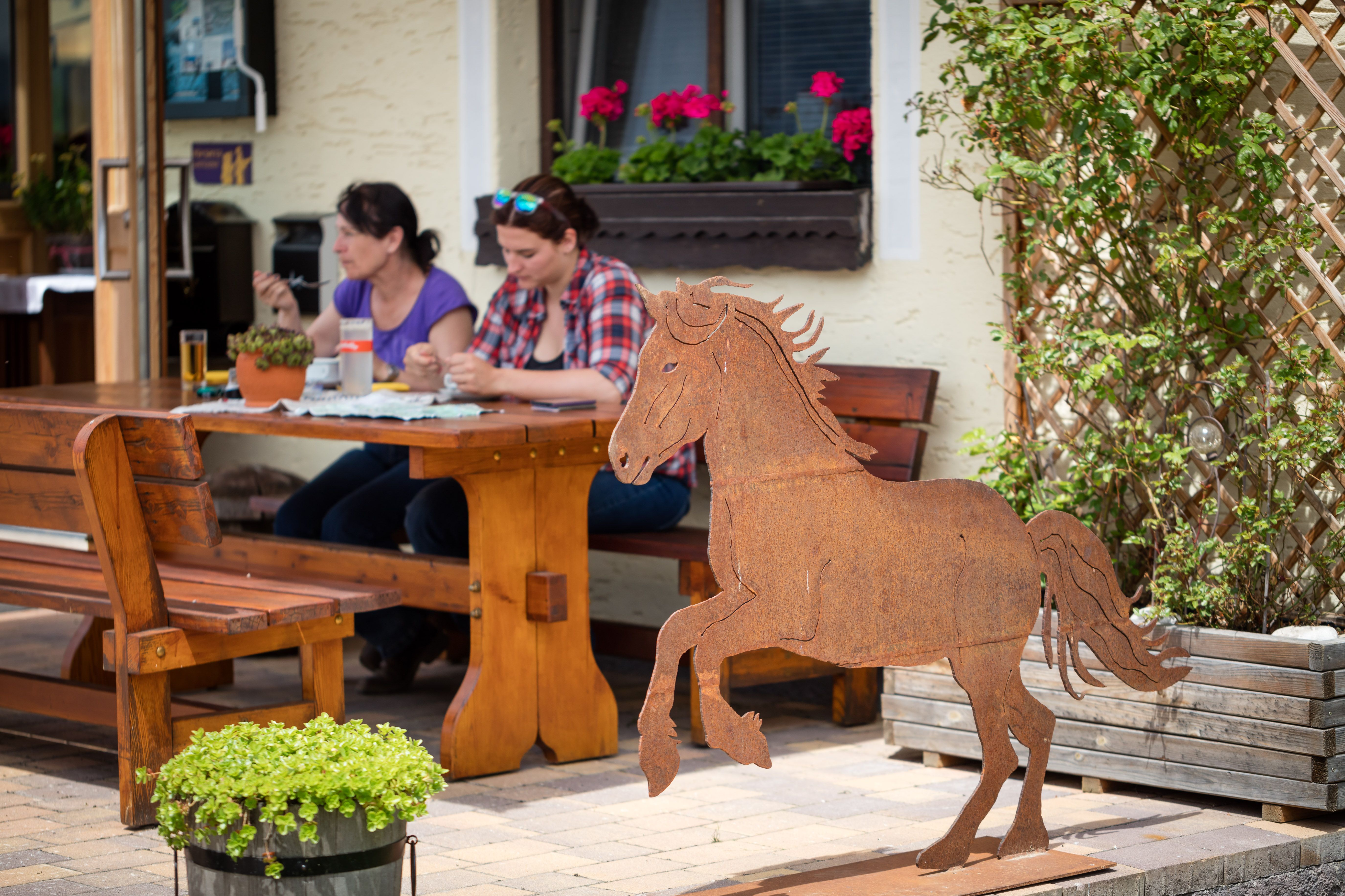 Zwei Frauen sitzen an einem Holztisch im Freien und essen. Im Vordergrund steht eine Metallskulptur eines Pferdes.