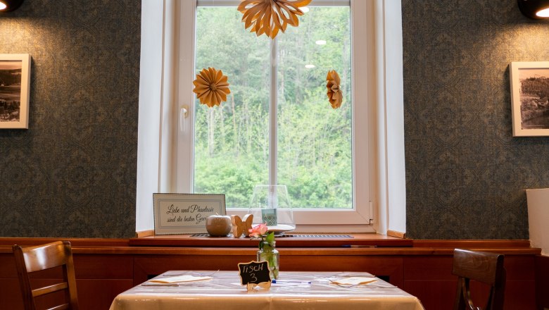 Ein Tisch in einem Restaurant mit Fensterblick, dekoriert mit Papierblumen und einem Schild mit der Aufschrift 'Liebe und Phantasie sind die besten Gewürze'.