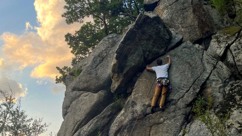 Person klettert an einer Felswand im Freien, umgeben von Bäumen und blauem Himmel mit Wolken.