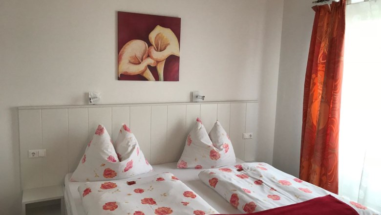 Schlafzimmer mit Doppelbett, Blumenbettw&auml;sche, rotem Bild und Vorh&auml;ngen.