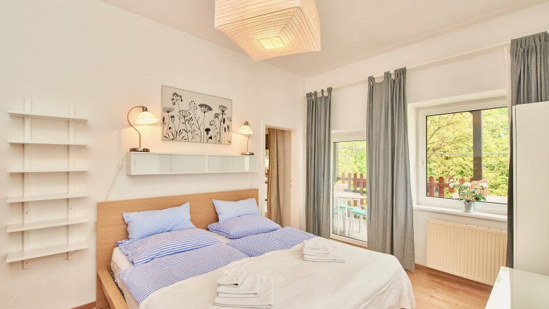 Helles Schlafzimmer mit Doppelbett, blauer Bettwäsche, zwei Lampen, einem Bild an der Wand und einem Fenster mit Blick auf einen Balkon.