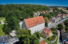 Luftaufnahme eines gro&szlig;en Geb&auml;udes mit rotem Dach, umgeben von B&auml;umen und kleineren H&auml;usern in einer l&auml;ndlichen Gegend.