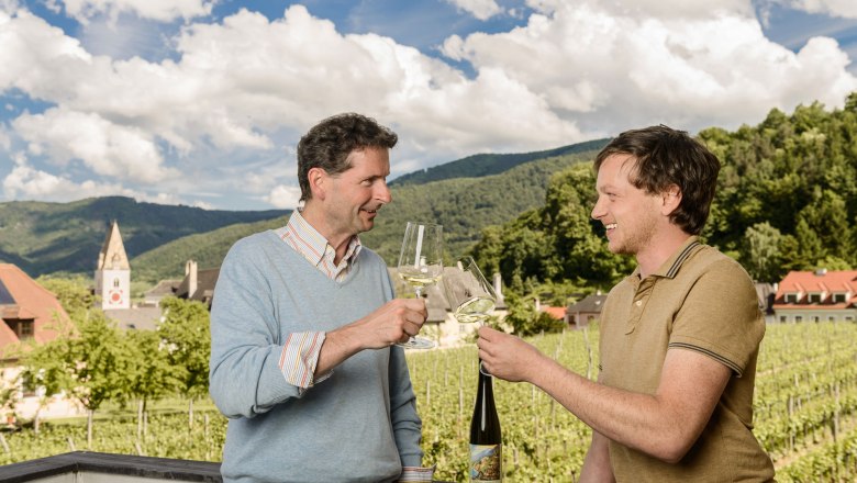 Zwei Männer stoßen mit Weingläsern an, im Hintergrund Weinberge und ein Kirchturm.