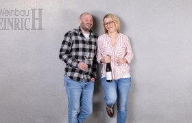Zwei Personen mit Weingläsern vor grauer Wand, Schriftzug 'Weinbau Heinrich'.