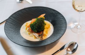 Ein elegant angerichtetes Gericht auf einem schwarzen Teller mit Gem&uuml;se und Sauce, daneben ein Glas Wei&szlig;wein auf einem gedeckten Tisch.