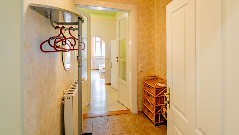 Ein Vorraum mit Kleiderhaken, einem Heizkörper und einem Korbmöbel. Eine Tür führt in einen hellen Raum mit Fenstern.