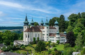 Ein wei&szlig;es Schloss mit mehreren Zwiebelt&uuml;rmen, umgeben von gr&uuml;ner Landschaft und B&auml;umen, unter einem blauen Himmel.