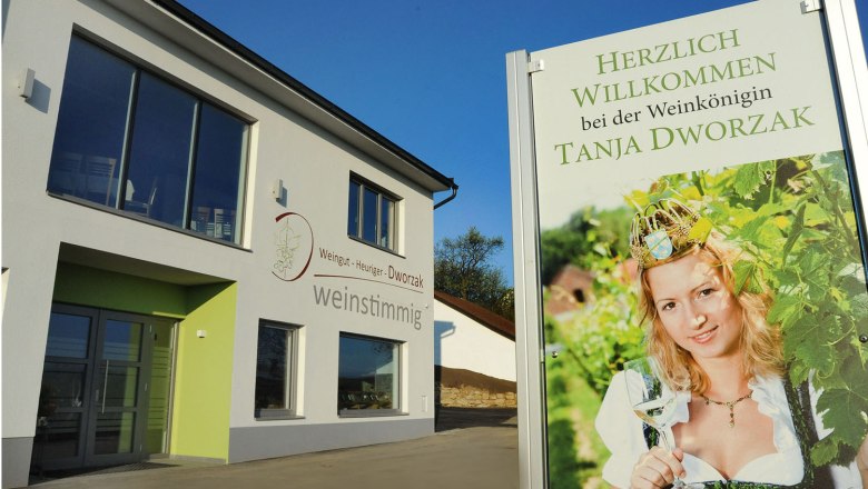 Ein modernes Geb&auml;ude mit einem Schild, das eine Frau in Tracht mit einem Weinglas zeigt. Text: Herzlich willkommen bei der Weink&ouml;nigin Tanja Dworzak.